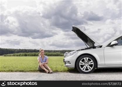Full length of worried businesswoman sitting by broken down car at countryside
