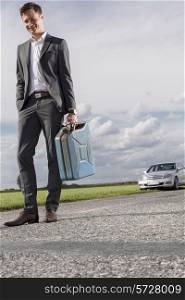 Full length of young businessman carrying gas can with broken down car in background at countryside