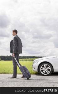 Full length of young businessman with suitcase leaving broken down car at countryside
