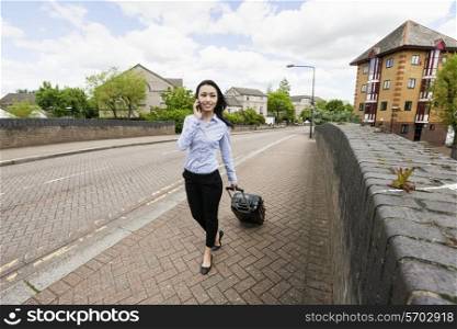 Full length of young businesswoman with luggage while answering cell phone walking on sidewalk