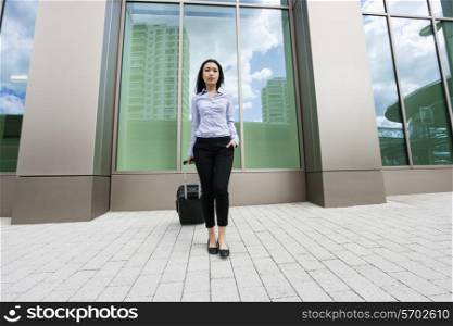 Full length of young businesswoman with suitcase on sidewalk against building