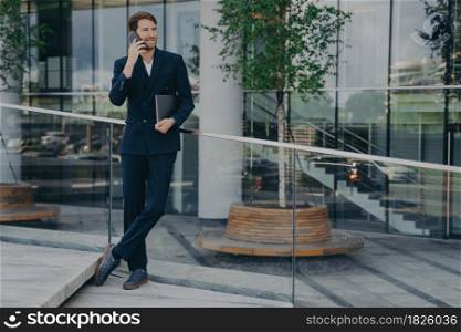 Full length of young confident man lawyer in stylish dark blue suit speaks on smartphone with smile while standing outside of business office center, holding laptop computer under his armpit. ?onfident man lawyer in dark blue suit speaks on smartphone with smile while standing outside
