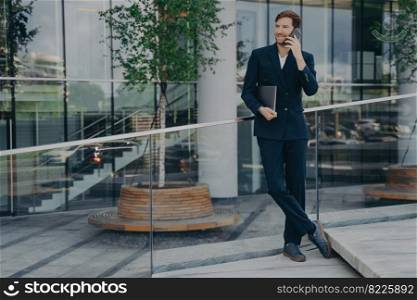 Full length of young confident man lawyer in stylish dark blue suit speaks on smartphone with smile while standing outside of business office center, holding laptop computer under his armpit.  onfident man lawyer in dark blue suit speaks on smartphone with smile while standing outside