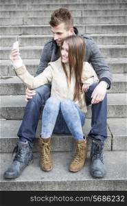 Full length of young couple taking picture of themselves while sitting on steps outdoors