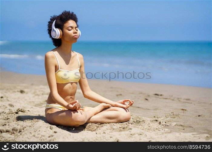 Full length of young fit African American lady with curly hair in bikini and headphones meditating in Padmasana, with closed eyes while practicing yoga on sandy beach. Tranquil black woman meditating in Lotus pose on sandy seacoast