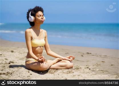 Full length of young fit African American lady with curly hair in bikini and headphones meditating in Padmasana, with closed eyes while practicing yoga on sandy beach. Tranquil black woman meditating in Lotus pose on sandy seacoast