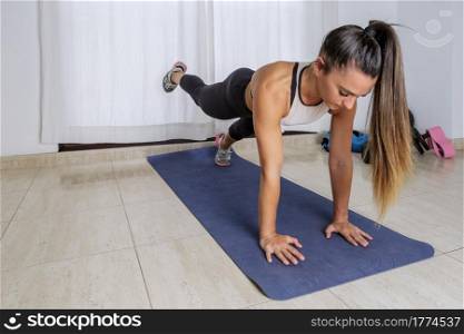 Full length of young fit female doing one legged plank exercise on mat during abs workout at home. Woman doing abs plank exercise