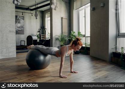 Full length of young hairy red haired woman doing exercises on fitball during workout in fitness studio, attractive female in active wear balancing on exercise ball and enjoying pilates training. Happy healthy woman balancing on exercise ball during fitness workout