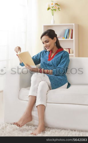 Full length of young Indian woman reading book on sofa