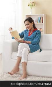 Full length of young Indian woman reading book on sofa