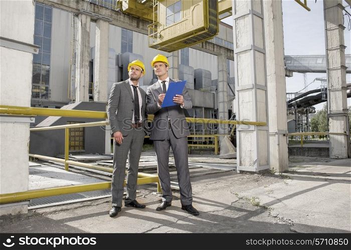 Full length of young male engineer with clipboard looking away at construction site