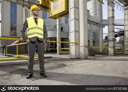 Full length of young male engineer with hands in pockets standing outside industry