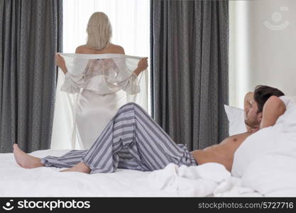 Full length of young man looking at woman removing robe at window