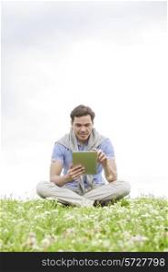 Full length of young man using digital tablet while sitting on grass against sky