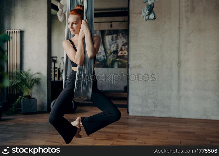 Full length of young slender woman with red hair practices anti gravity fly yoga inside hammock, happy female doing relaxing exercises in fitness studio. Concept of healthy lifestyle and sport indoor. Slender young woman with red hair practices anti-gravity fly yoga