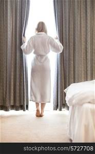 Full length of young woman in bathrobe opening bedroom curtains at hotel room