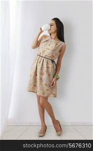 Full length of young woman in sundress drinking coffee against white wall