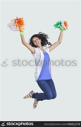 Full length of young woman jumping in mid-air with Indian tricolor pom-poms over colored background