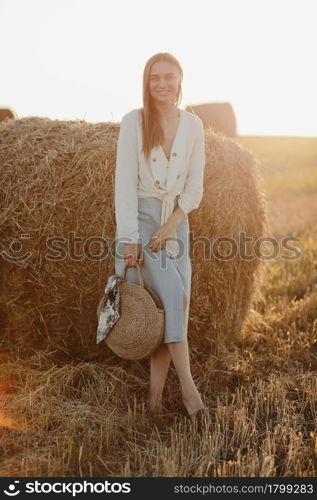 Full length portrait of a smiling beautiful brunette in a jeans skirt and straw bag in hand. Woman enjoying a walk in a wheat field with hay bales on summer sunny day. Full length portrait of a smiling beautiful brunette in a jeans skirt and straw bag in hand. Woman enjoying a walk in a wheat field with hay bales on summer sunny day.