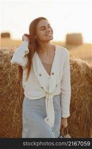 Full length portrait of a smiling beautiful brunette in a jeans skirt and straw bag in hand. Woman enjoying a walk in a wheat field with hay bales on summer sunny day. Full length portrait of a smiling beautiful brunette in a jeans skirt and straw bag in hand. Woman enjoying a walk in a wheat field with hay bales on summer sunny day.