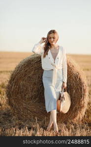 Full length portrait of a smiling beautiful brunette in a jeans skirt and straw bag in hand. Woman enjoying a walk in a wheat field with hay bales on summer sunny day. Full length portrait of a smiling beautiful brunette in a jeans skirt and straw bag in hand. Woman enjoying a walk in a wheat field with hay bales on summer sunny day.