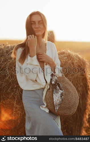 Full length portrait of a smiling beautiful brunette in a jeans skirt and straw bag in hand. Woman enjoying a walk in a wheat field with hay bales on summer sunny day. Full length portrait of a smiling beautiful brunette in a jeans skirt and straw bag in hand. Woman enjoying a walk in a wheat field with hay bales on summer sunny day.