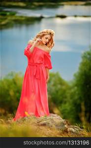 Full length portrait of beautiful sensual young blond woman in long pink dress and floral wreath near the lake outdoors in natural background. Full length portrait of beautiful sensual young blond woman in long pink dress and floral wreath near the lake outdoors in natural background.