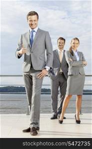Full length portrait of confident businessman gesturing thumbs up while standing with coworkers on terrace against sky