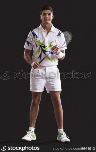 Full length portrait of confident young man with tennis racket standing against black background