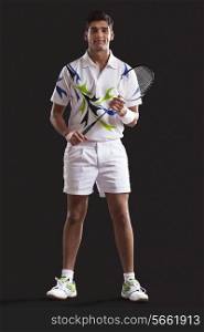 Full length portrait of confident young man with tennis racket standing against black background