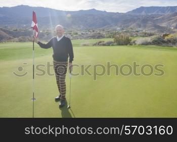 Full length portrait of happy senior male golfer holding flag and putter at golf course