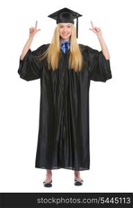 Full length portrait of happy young woman in graduation gown pointing up on copy space