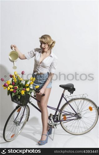 full-length portrait of sensual blonde woman with denim shorts on the bicycle watering coloured tulips in the basket