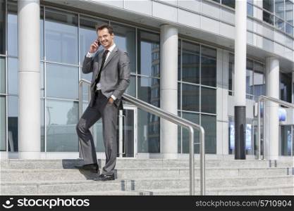 Full length portrait of smiling businessman answering cell phone while standing on steps outside office