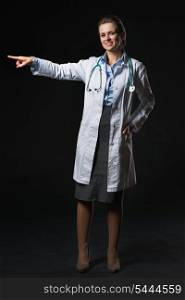 Full length portrait of smiling doctor woman pointing on copy space isolated on black