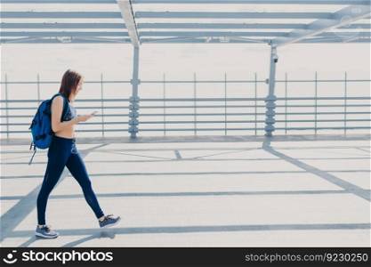 Full length portrait of sporty active female teenager or youngster, wears top, leggings and sneakers, carries rucksack, enjoys pleasant music in playlist, uses modern cell phone and earphones