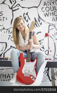 Full-length portrait of teenage girl with electric guitar sitting on study table at home