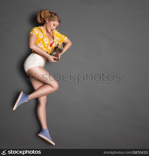 Full-length profile of a beautiful pin-up girl, looking at an old vintage photo camera with bulb flash on chalkboard background.