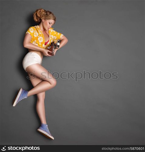 Full-length profile of a beautiful pin-up girl, looking at an old vintage photo camera with bulb flash on chalkboard background.