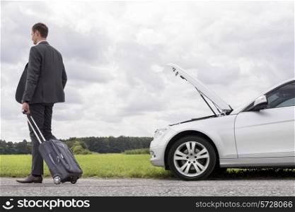 Full length rear view of young businessman with luggage leaving broken down car at countryside