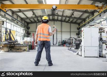 Full length rear view of young manual worker operating crane in factory