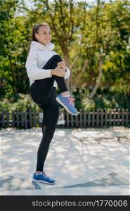 Full length shot of active healthy young woman warms up before jogging in park keeps knee raised wears sweatshirt leggings and sneakers listens music in earphones. Healthy lifestyle concept.