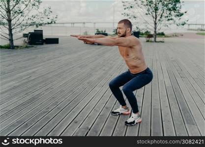 Full length shot of active man does squatting exercise poses with naked torso has muscular body poses outside. Strong athletic fit man in sportswear exercises in open air. Confident fitness instructor