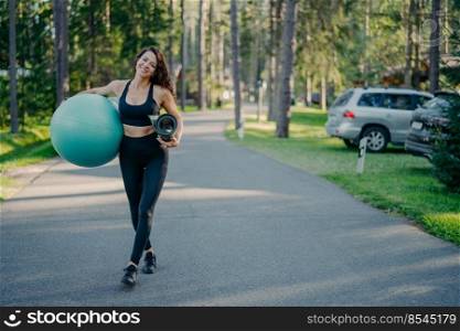 Full length shot of active slim woman carries fitness ball and rolled up karemat, dressed in leggings, top and sneakers, walks on path near park or forest, looks gladfully, has athletic body