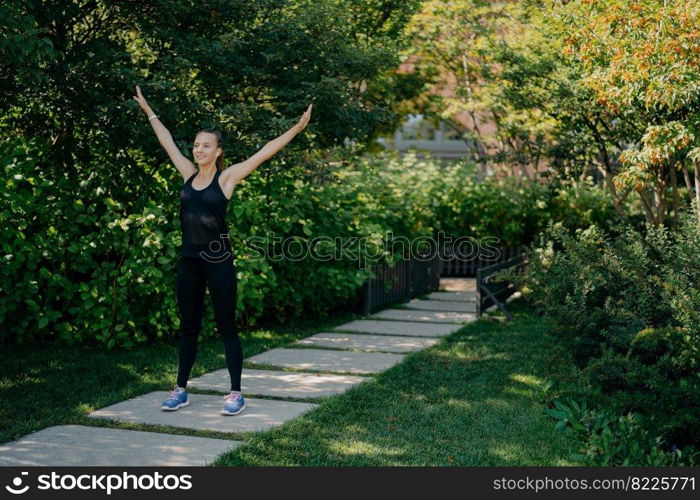 Full length shot of active sportswoman raises arms breathes deeply dressed in active wear has sport training in park during morning time listens music in earphones. Sporty lifestyle concept.