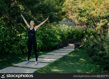 Full length shot of active sportswoman raises arms breathes deeply dressed in active wear has sport training in park during morning time listens music in earphones. Sporty lifestyle concept.