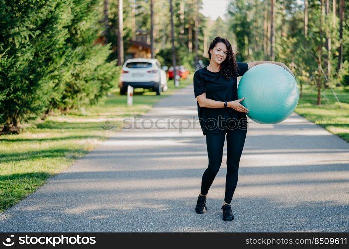 Full length shot of active sporty woman holds big fitness ball exercises outdoor walks on road near forest leads healthy lifestyle stays fit dressed in sportswear. People gymnastics and aerobics