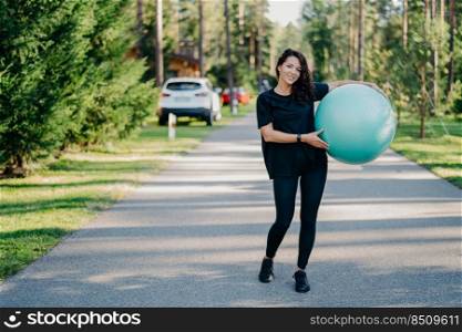 Full length shot of active sporty woman holds big fitness ball exercises outdoor walks on road near forest leads healthy lifestyle stays fit dressed in sportswear. People gymnastics and aerobics