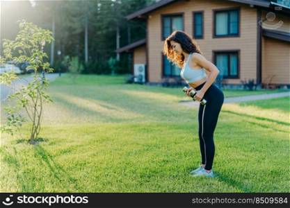 Full length shot of active sporty young woman raises dumbbells has workout outdoor near house stands on green lawn, dressed in sportsclothes, tries to be in good shape, stays fit and healthy
