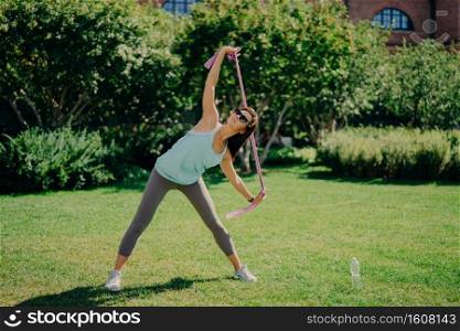 Full length shot of active woman stretches arms with fitness gum has good flexibility wears t shirt leggings and sneakers has workout with resistance band poses outdoor on green lawn during summer